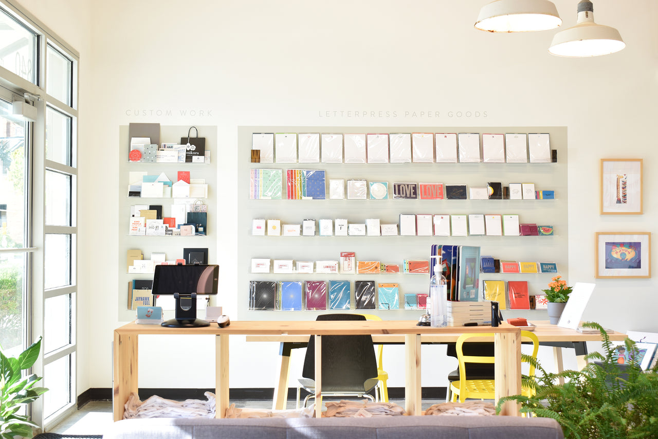 The Inkello Studio shop area with rows of stationery, greeting cards and printed samples. There is a big meeting table, some green plants, and framed illustrations hanging on the wall. The photo was taken on a bright and sunny day.
