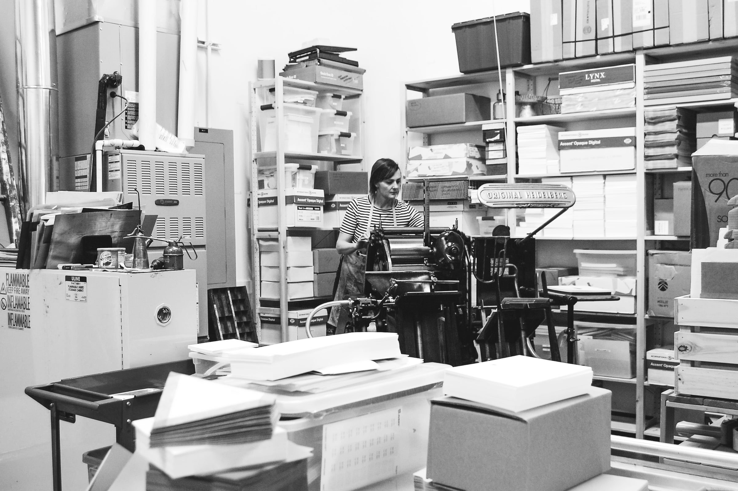 Christy is printing greeting cards on the large Windmill printing press. There are shelves and stacks of paper around the corner of the room.
